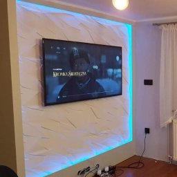 Kompleksowe Remonty - Panel 3D z podświetleniem LED za telewizorem, nowoczesne wykończenie ściany w salonie. Białe panele o geometrycznym wzorze. Ciepłe światło z góry, ciemna podłoga.