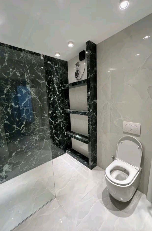 Elegancka łazienka z kabiną prysznicową wykończoną czarnym marmurem i białą toaletą, podkreślona oświetleniem punktowym i półkami w ścianie.