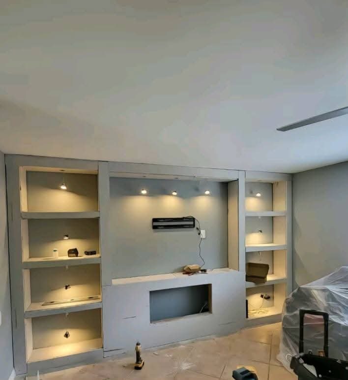 Ściana działowa z regipsu z wnękami na półki i TV w szarym odcieniu, oświetlenie punktowe, w trakcie prac wykończeniowych. Widoczne narzędzia pracy.