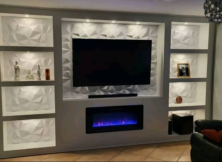 Stylowa ściana TV z geometrycznymi panelami 3D i elektrycznym kominkiem, oświetlona punktowo. Dekoracyjne figurki i ramka na półkach wnękowych.
