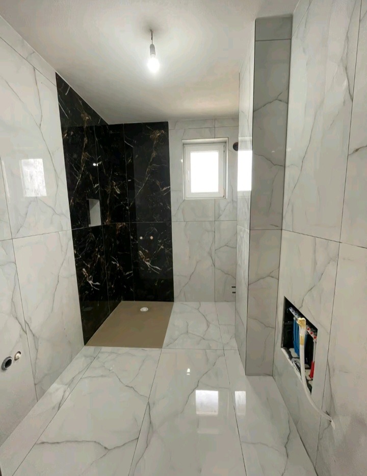 Wykończona łazienka z czarno-białymi płytkami imitującymi marmur, widoczny brodzik i okno. W ścianie otwór na instalacje. Całość w jasnej tonacji.