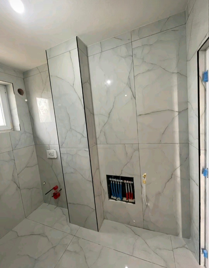 Wykończona łazienka z marmurowymi płytkami, widoczne przyłącza hydrauliczne w ścianie i podwieszana toaleta. Minimalistyczny design, szare żyły na białym tle płytek.