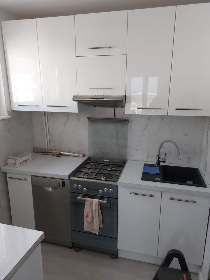 Biała, funkcjonalna kuchnia z połyskiem: zabudowa z szafkami górnymi i dolnymi, kuchenka gazowa, zlewozmywak w blacie. Marmurowe wykończenie ściany.