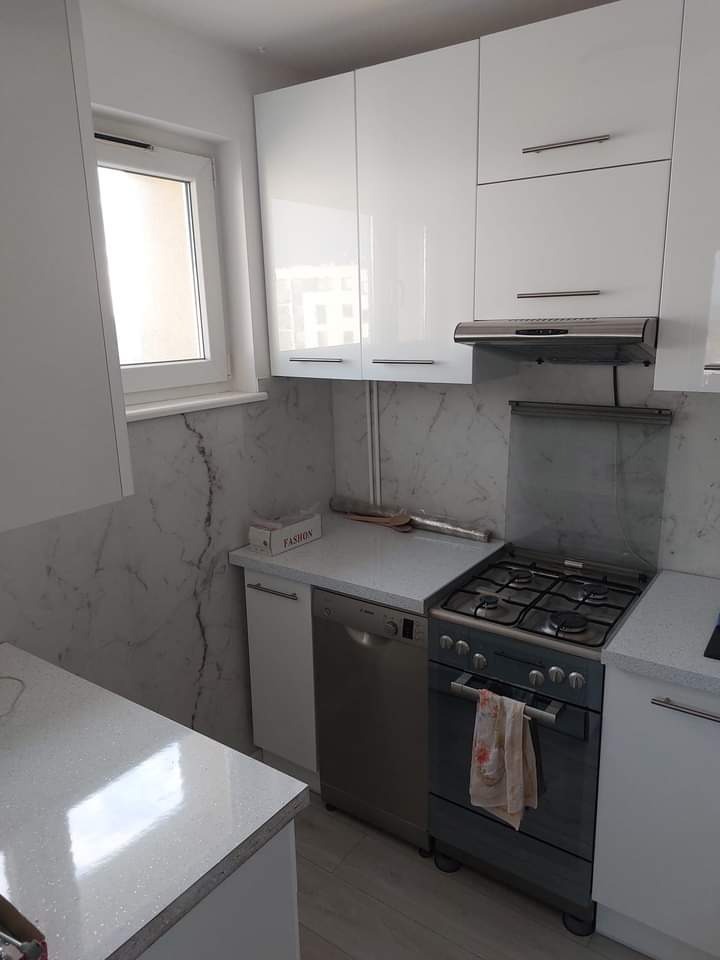 Nowoczesna, jasna kuchnia po remoncie: białe, błyszczące szafki, marmurowe płytki na ścianie, zabudowana zmywarka i kuchenka gazowa. Okno z widokiem na zewnątrz.