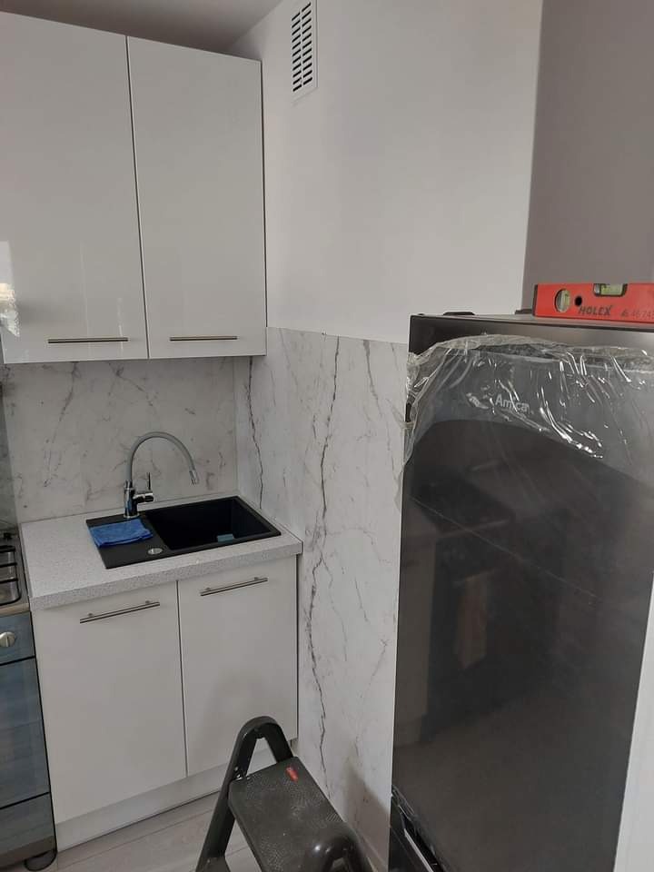 Nowoczesna kuchnia po remoncie: białe szafki z marmurowym blatem i ścianą, czarny zlew, nowy sprzęt AGD w folii. Poziomica na lodówce.