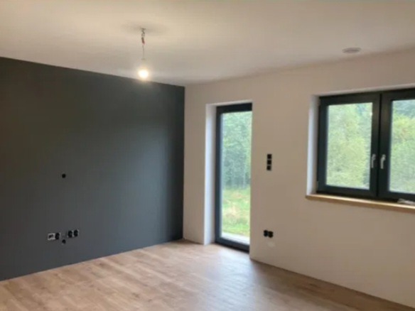 Wnętrze pokoju po malowaniu: jedna ściana grafitowa, reszta biała. Widoczne okno z czarną ramą, drewniana podłoga i przygotowane punkty elektryczne.