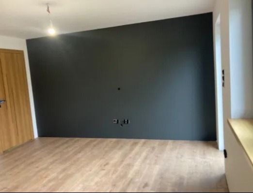 Pomalowane na ciemny kolor ściany w pokoju z drewnianą podłogą i drzwiami. Widoczne gniazdka elektryczne na ścianie i wiszący kabel od lampy na suficie.