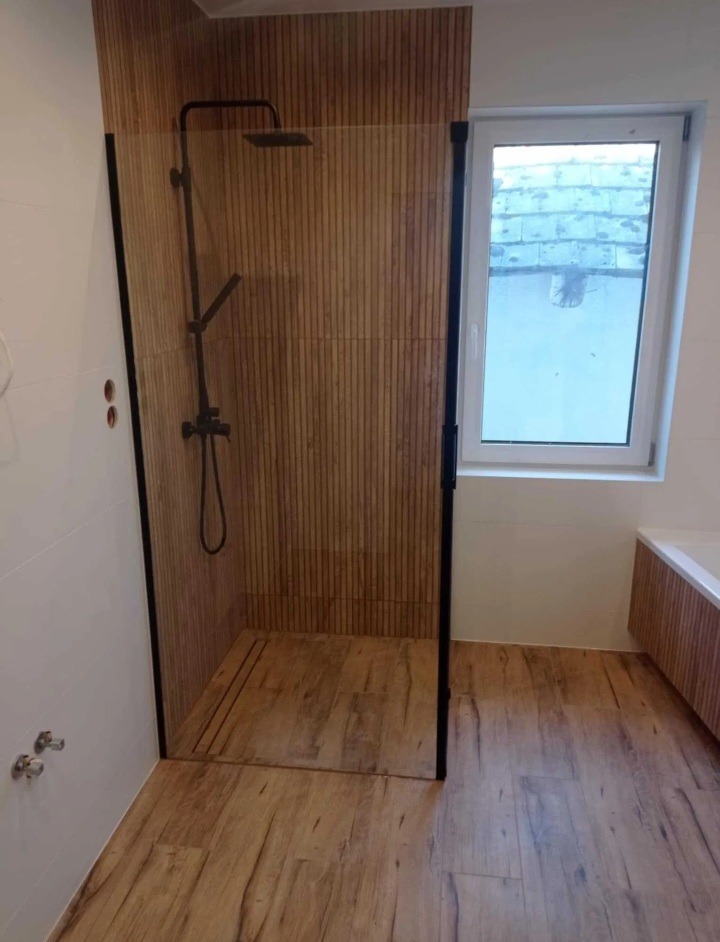 Nowoczesna kabina prysznicowa z czarnymi ramami i deszczownicą, drewnopodobne płytki na ścianach i podłodze, okno z widokiem na dach.