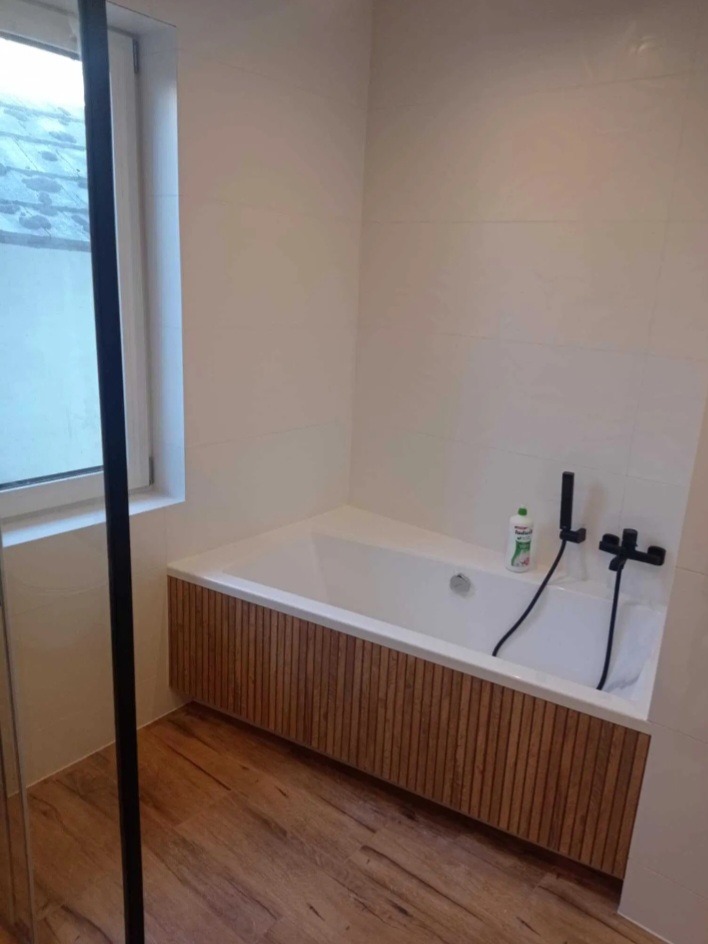 Nowoczesna łazienka z białą wanną obudowaną drewnianymi lamelami, drewnopodobną podłogą i białą glazurą. Czarna armatura i okno z widokiem na zewnątrz.