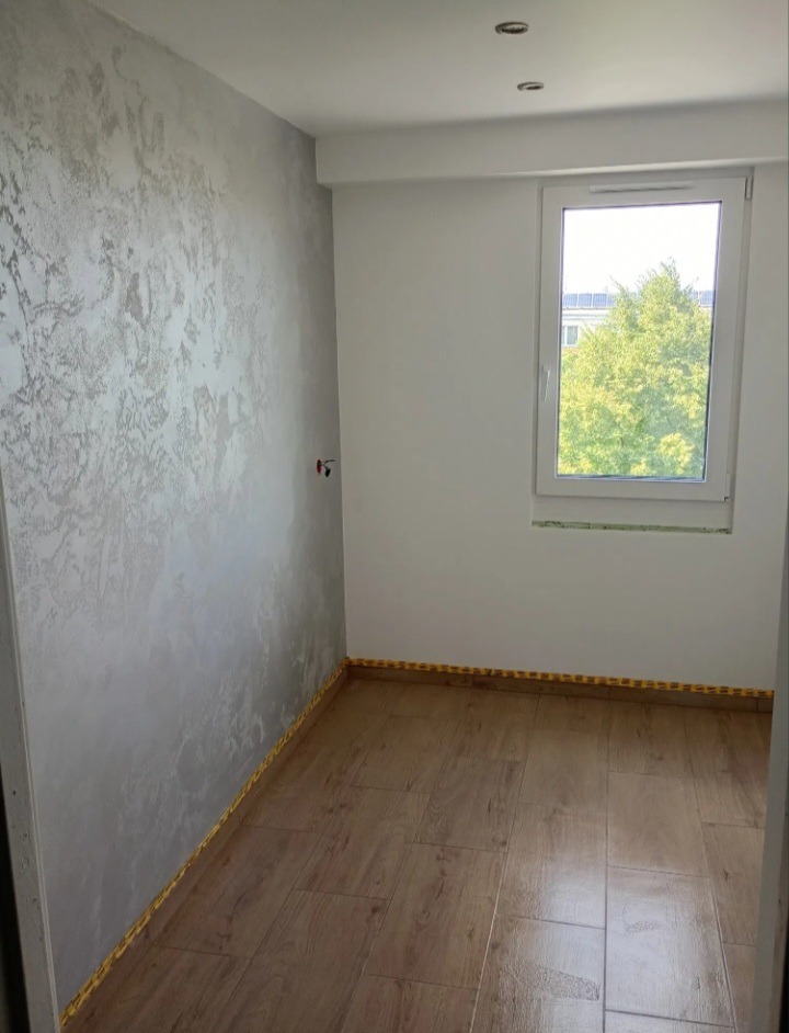 Pokój po remoncie: gładź dekoracyjna w odcieniach szarości na ścianie, parkiet imitujący drewno, okno z widokiem na zieleń. Wykończone listwy przypodłogowe.