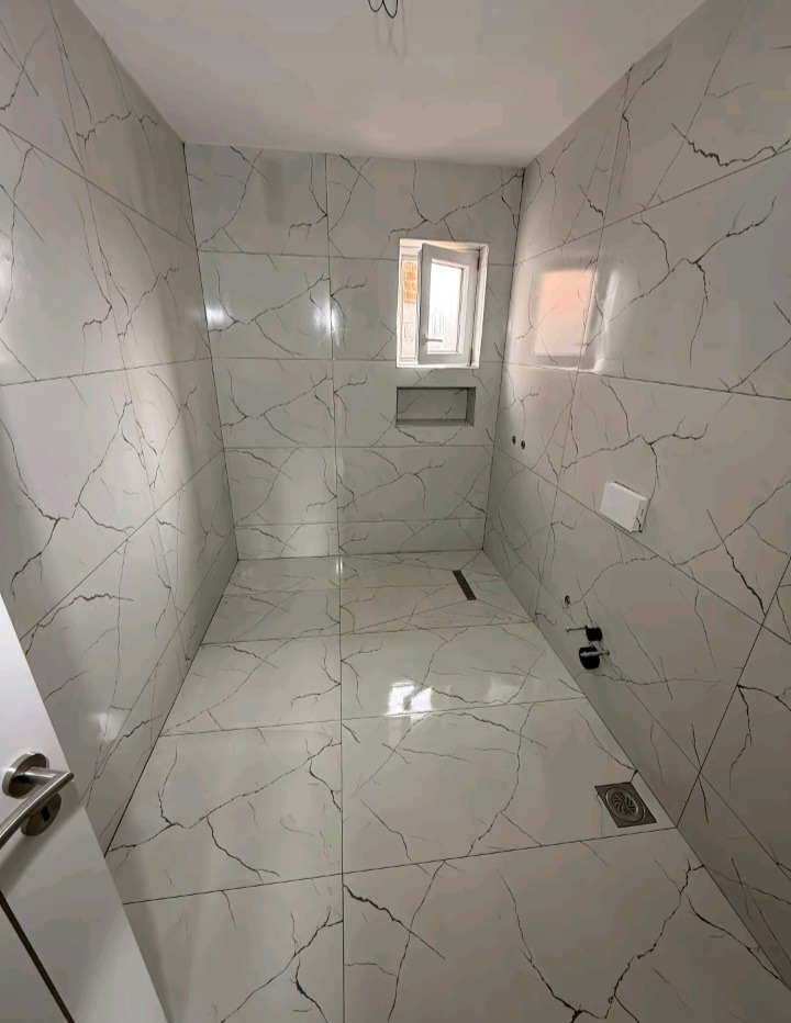 Wykończona łazienka z białymi płytkami imitującymi marmur, małe okno z wnęką i odpływy liniowe. Widok na całe pomieszczenie z perspektywy wejścia.