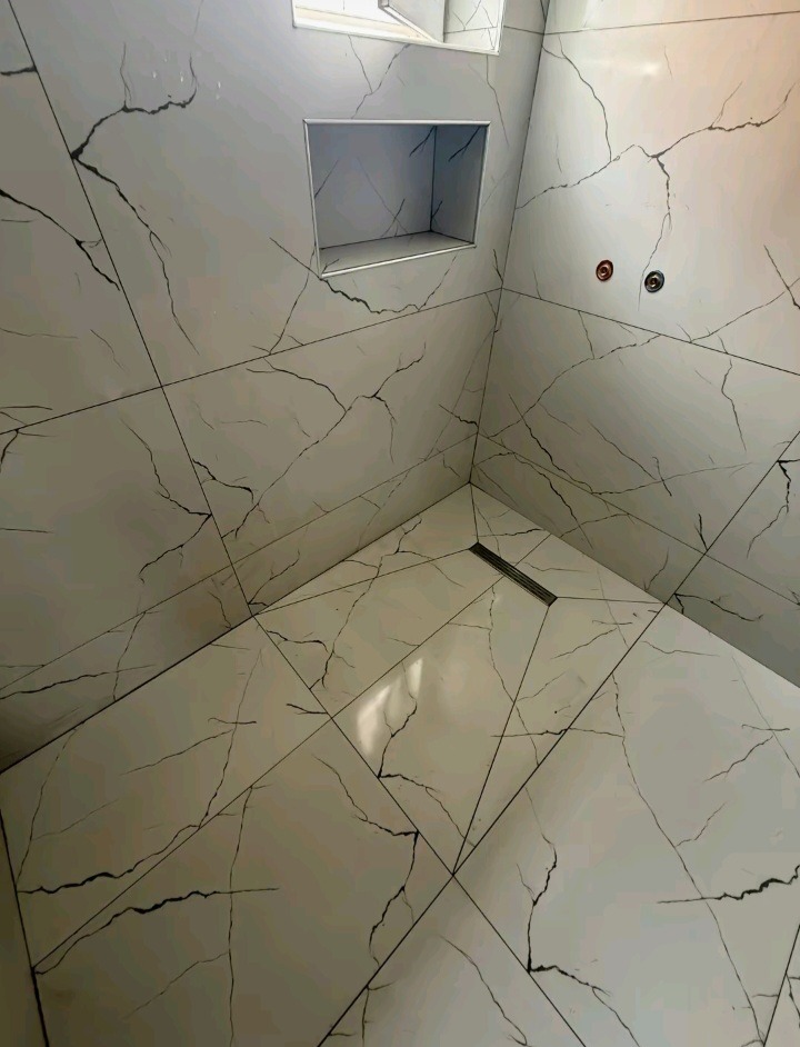 Wykończona kabina prysznicowa z białymi płytkami imitującymi marmur, z wnęką i odpływem liniowym. Minimalistyczny design, nowoczesne wykończenie.