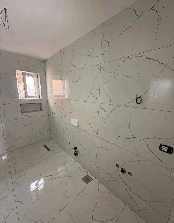 Wykończona łazienka z białymi płytkami imitującymi marmur, widoczne odpływy liniowe i punkty instalacji wodnej. Małe okno z wnęką. Nowoczesny design.