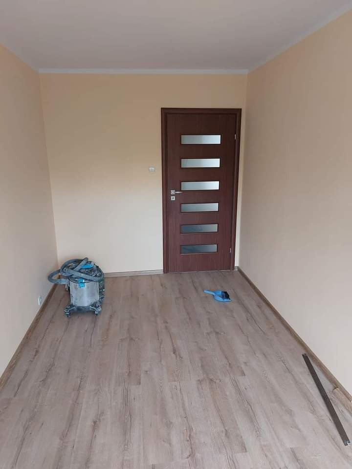 Jasny pokój po remoncie: świeżo pomalowane ściany w odcieniach beżu, brązowe drzwi z przeszkleniami i panele podłogowe. Widoczny odkurzacz i listwa przypodłogowa.