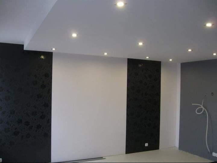 Wykończone wnętrze pokoju z białymi i czarnymi panelami ściennymi, oświetleniem punktowym sufitowym i szarą ścianą z widocznymi przewodami.