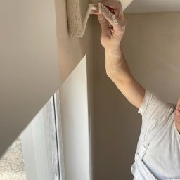 CraftGips - Pracownik z wałkiem maluje ścianę w Żabiej Woli. Widoczny fragment okna i taśma malarska. Ręka pracownika pokryta farbą.