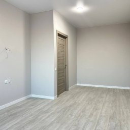 CraftGips - Jasne, puste wnętrze po remoncie: szare ściany, drewnopodobna podłoga, minimalistyczne listwy przypodłogowe i widoczne gniazdko elektryczne. Drzwi wewnętrzne w odcieniach szarości.