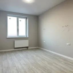 Jasne, puste wnętrze pokoju po remoncie. Szare ściany, białe okno i grzejnik. Podłoga wyłożona panelami imitującymi drewno. Widoczne wystające kable elektryczne.