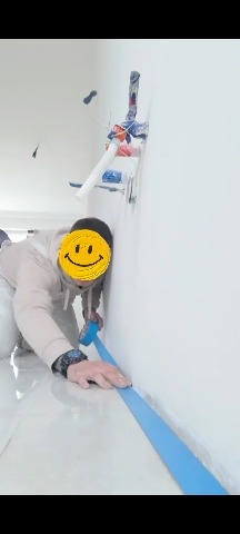 Pracownik zabezpiecza taśmą podłogę przy ścianie przed malowaniem. Widoczne białe płytki podłogowe i fragment instalacji na ścianie.