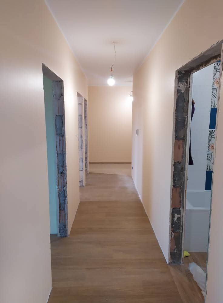 Korytarz po malowaniu ścian na delikatny beż, widoczne ościeżnice drzwiowe przed montażem, podłoga wyłożona panelami imitującymi drewno.
