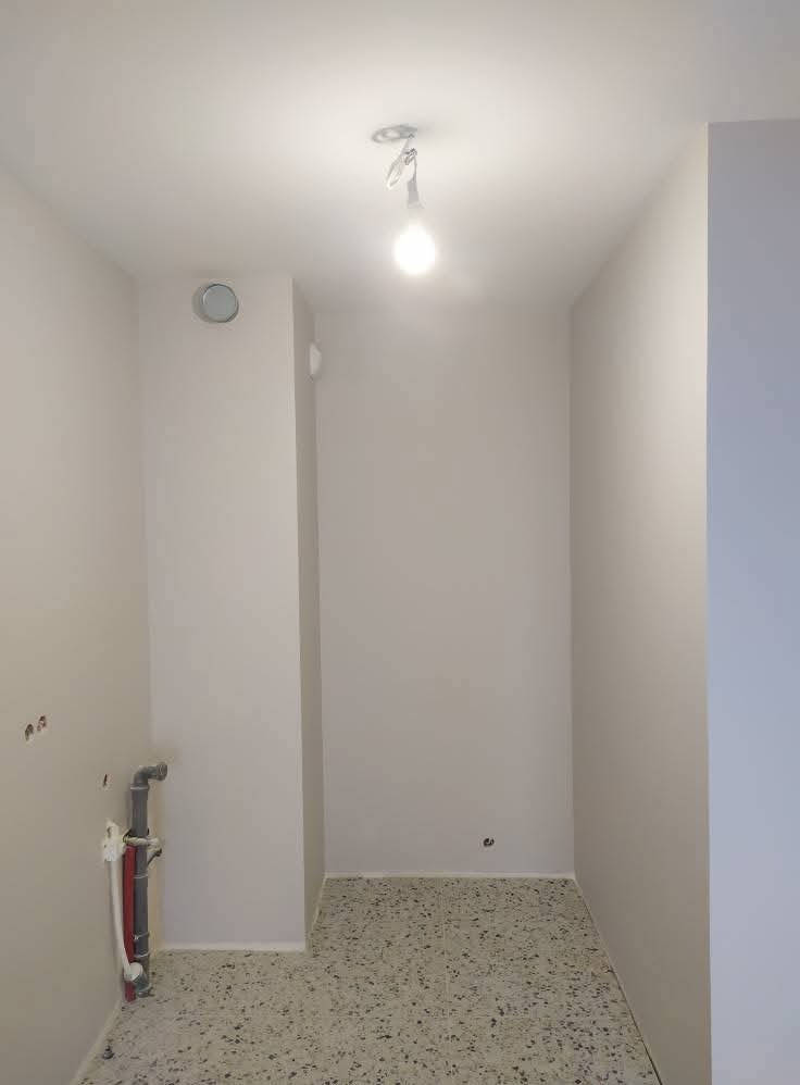 Wnętrze po remoncie: gładkie, białe ściany, sufit z żarówką i podłoga z terazzo. Widoczne rury przy ścianie. Minimalistyczne wykończenie.