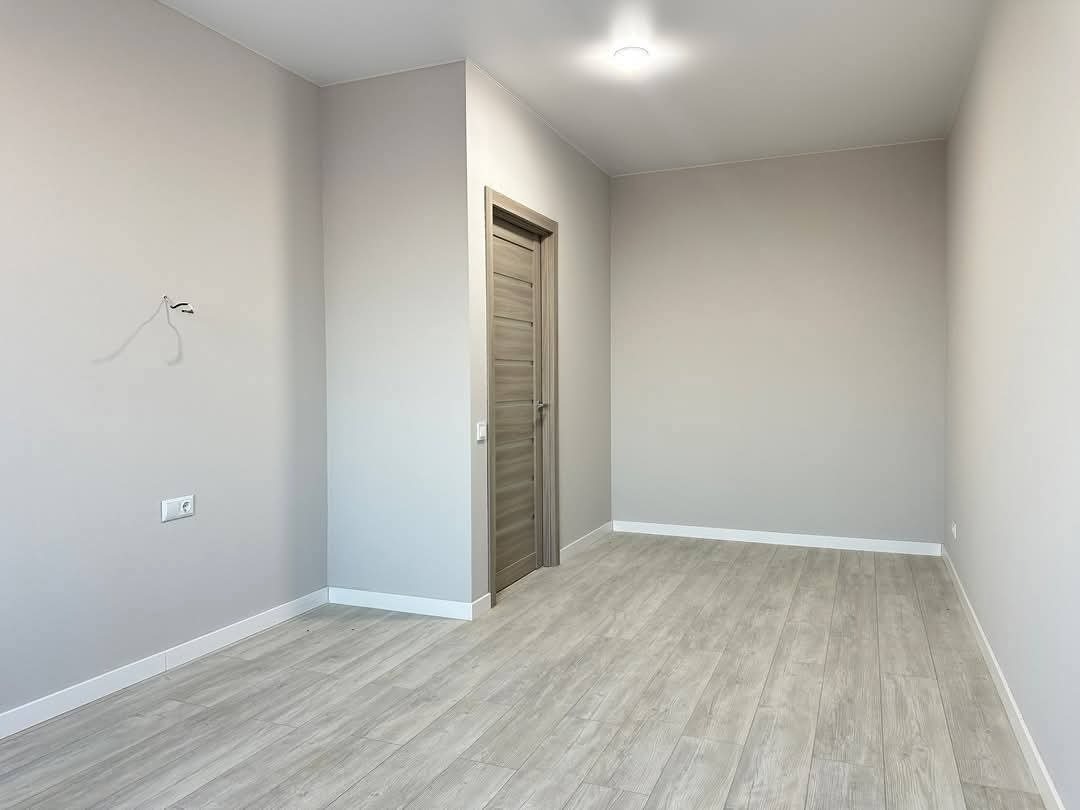 Jasne, puste wnętrze po remoncie: szare ściany, drewnopodobna podłoga, minimalistyczne listwy przypodłogowe i widoczne gniazdko elektryczne. Drzwi wewnętrzne w odcieniach szarości.