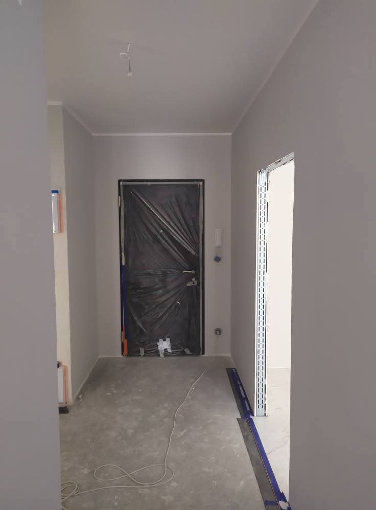 Korytarz po szpachlowaniu i malowaniu na szaro, drzwi zabezpieczone folią, widoczne profile do wykończenia ościeżnicy, przygotowany do dalszych prac.