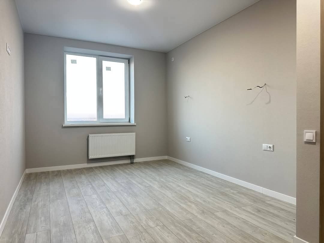 Jasne, puste wnętrze pokoju po remoncie. Szare ściany, białe okno i grzejnik. Podłoga wyłożona panelami imitującymi drewno. Widoczne wystające kable elektryczne.