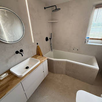 Nowoczesna łazienka z wanną asymetryczną, umywalką nablatową na drewnianym blacie i okrągłym lustrem. Minimalistyczny design, szare płytki, czarna armatura.