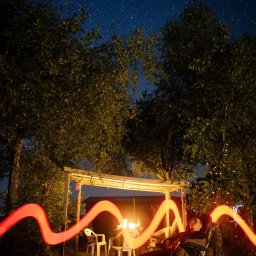 PR Graphics Zumkowski-Pawłowicz - Nocne ujęcie relaksującego mężczyzny w ogrodzie, oświetlonego długą ekspozycją czerwonej smugi światła, z widokiem na gwiazdy i altanę z lampkami.