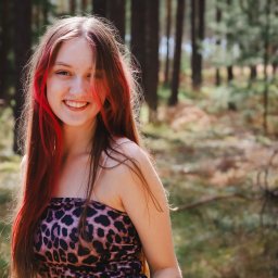 PR Graphics Zumkowski-Pawłowicz - Uśmiechnięta nastolatka z długimi, brązowymi włosami z czerwonymi pasemkami, w bluzce w panterkę, pozuje na tle zamazanego, leśnego krajobrazu.