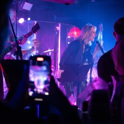 PR Graphics Zumkowski-Pawłowicz - Wokalistka śpiewająca do mikrofonu na scenie podczas koncertu, oświetlona fioletowo-niebieskim światłem, z gitarzystą i publicznością nagrywającą telefonami.
