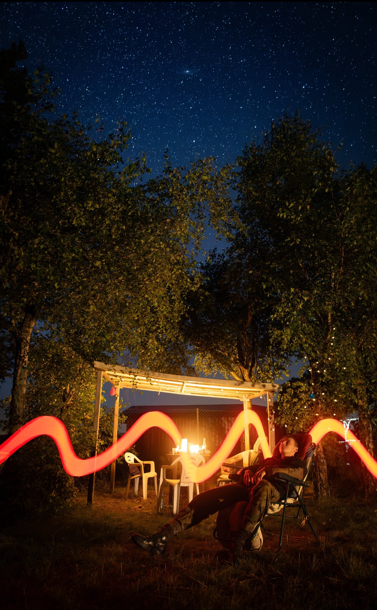 Nocne ujęcie relaksującego mężczyzny w ogrodzie, oświetlonego długą ekspozycją czerwonej smugi światła, z widokiem na gwiazdy i altanę z lampkami.