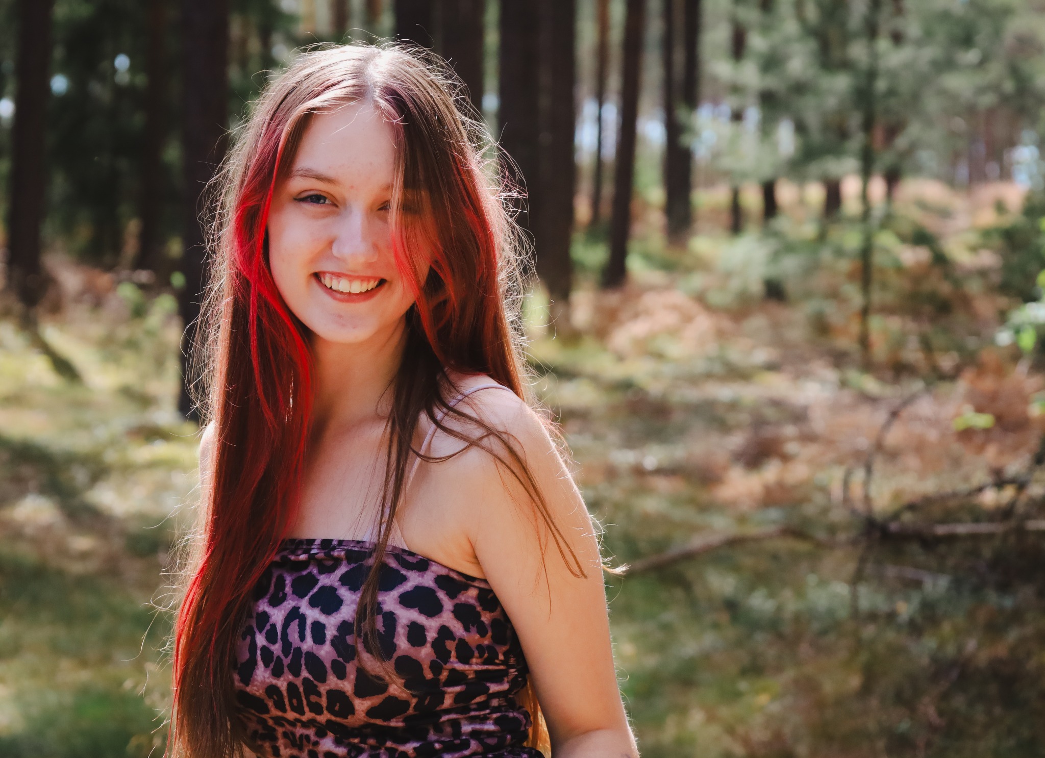 Uśmiechnięta nastolatka z długimi, brązowymi włosami z czerwonymi pasemkami, w bluzce w panterkę, pozuje na tle zamazanego, leśnego krajobrazu.