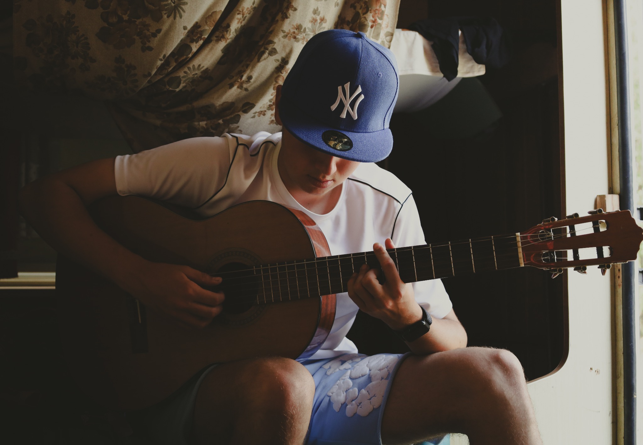 Młody mężczyzna w czapce z daszkiem gra na gitarze klasycznej w pomieszczeniu z wzorzystą zasłoną w tle. Skupienie na dłoniach i gryfie gitary.