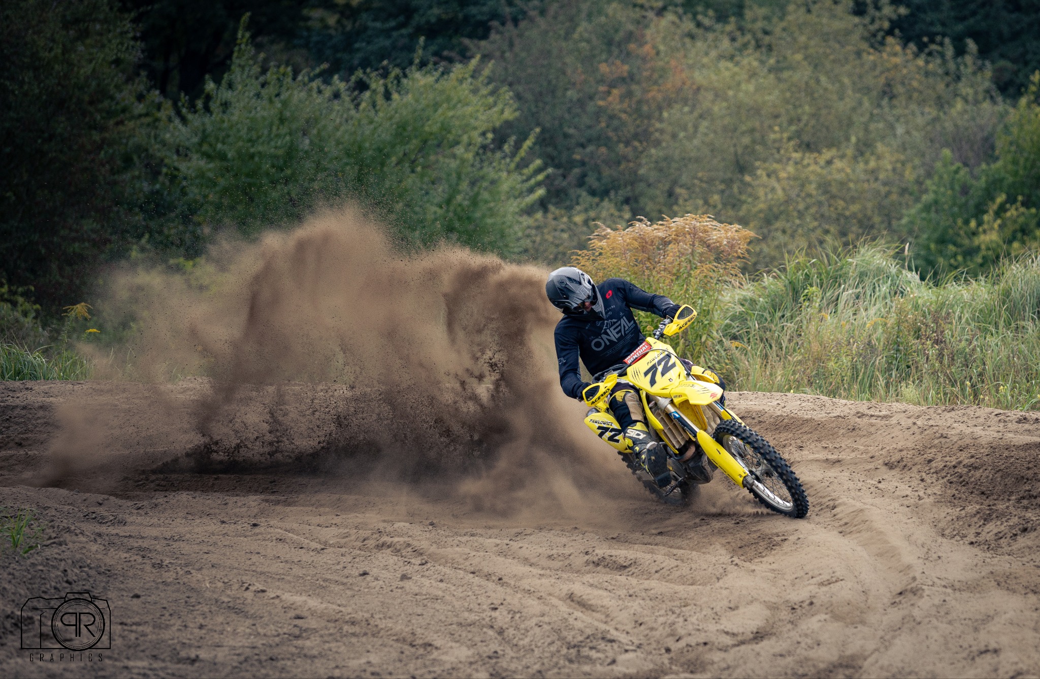 Żółty motocykl crossowy w akcji, dynamiczne ujęcie z piaskiem w tle, zawodnik w czarnym stroju i kasku, numer 72 na maszynie. Widoczne logo fotografa.