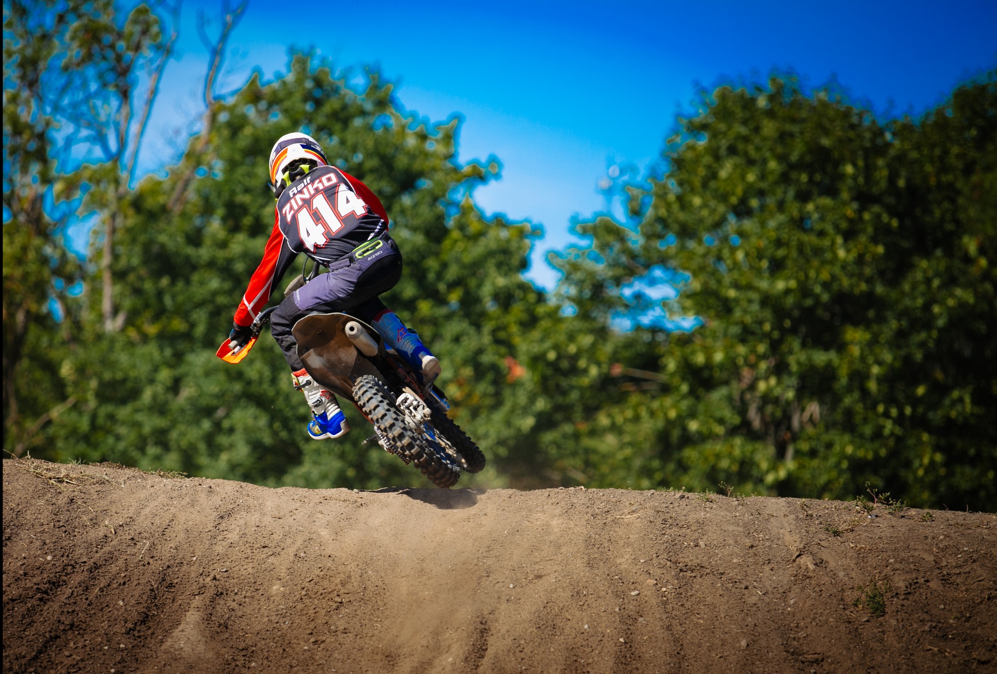 Dynamiczne ujęcie motocyklisty w locie nad ziemnym wzniesieniem, z numerem 414 na plecach, na tle zieleni i błękitnego nieba.