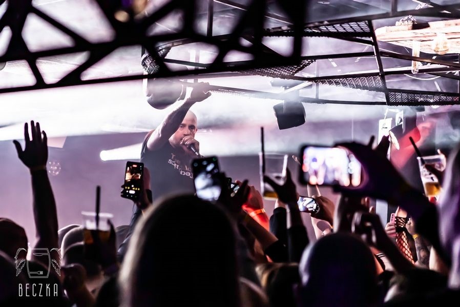 Koncert w klubie: raper na scenie, oświetlenie, tłum fanów nagrywających występ telefonami. Widoczny fragment konstrukcji sufitu i logo klubu 'Beczka'.