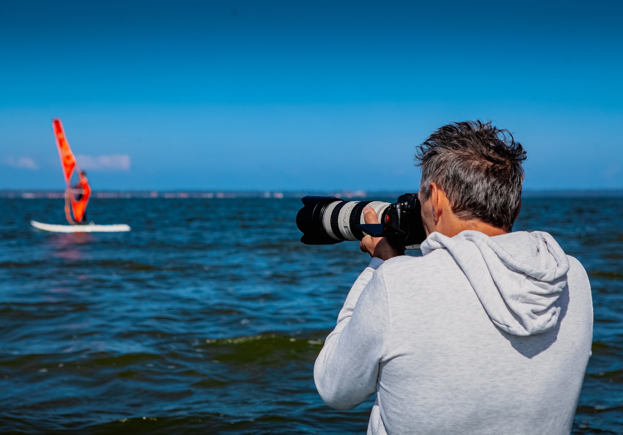 Fotograf z teleobiektywem robi zdjęcie windsurfera na morzu w słoneczny dzień. Ujęcie od tyłu, z widoczną sylwetką sportowca w oddali.