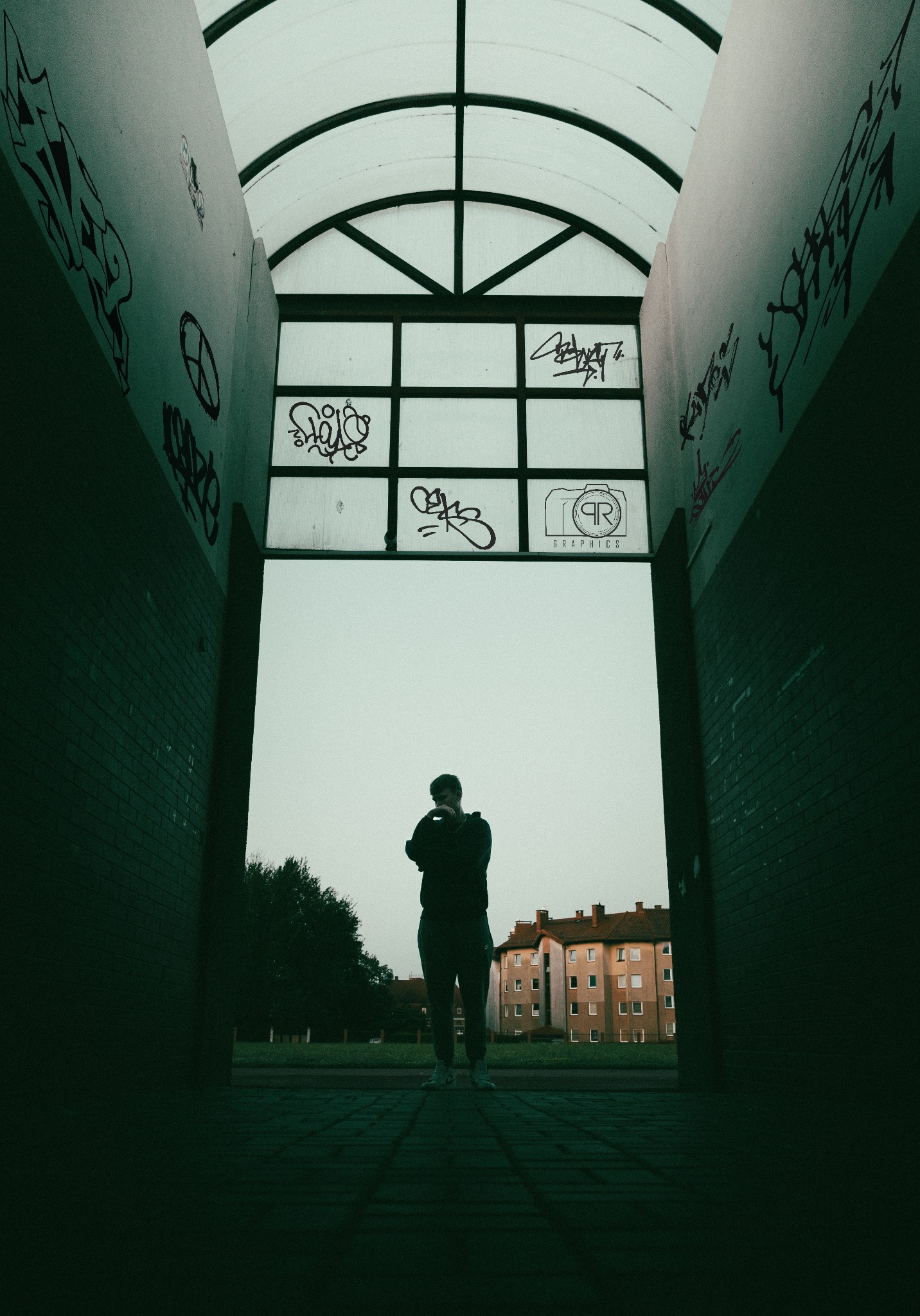 Sylwetka mężczyzny w tunelu z graffiti i logo 'QR Graphics' na szybie. Budynek mieszkalny w tle, pochmurne niebo.