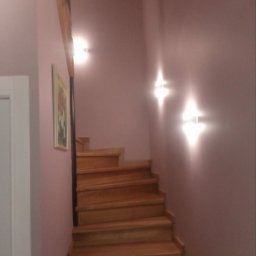 DMbudd Vadym Rak - Drewniane schody w odcieniach brązu, oświetlone lampami ściennymi, na tle ściany w kolorze pudrowego różu z obrazem w ramie.