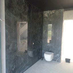 DMbudd Vadym Rak - Wykończona łazienka z czarnym sufitem i ścianami imitującymi marmur. Widoczne białe sanitariaty i szara podłoga. W tle widoczny remontowany pokój.
