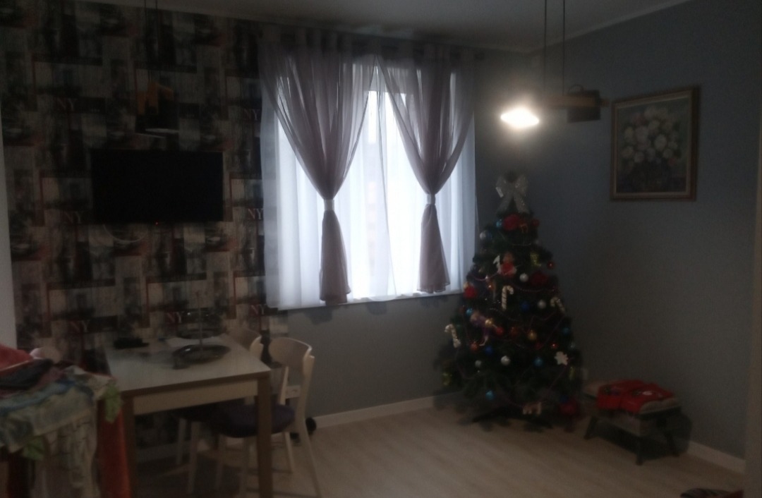 Udekorowane wnętrze pokoju z tapetą w stylu nowojorskim, firanami i choinką. W tle stół z krzesłami, lampa i obraz.