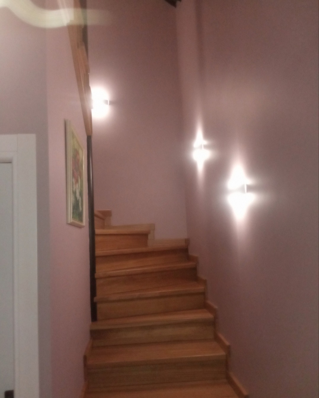 Drewniane schody w odcieniach brązu, oświetlone lampami ściennymi, na tle ściany w kolorze pudrowego różu z obrazem w ramie.