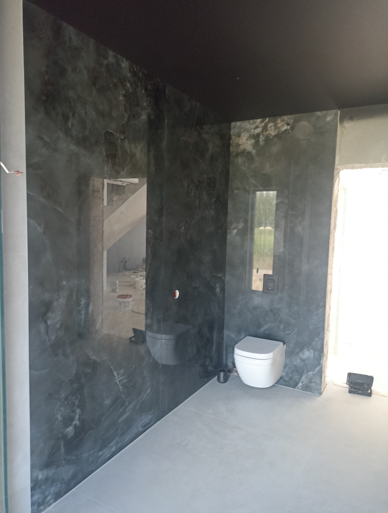 Wykończona łazienka z czarnym sufitem i ścianami imitującymi marmur. Widoczne białe sanitariaty i szara podłoga. W tle widoczny remontowany pokój.