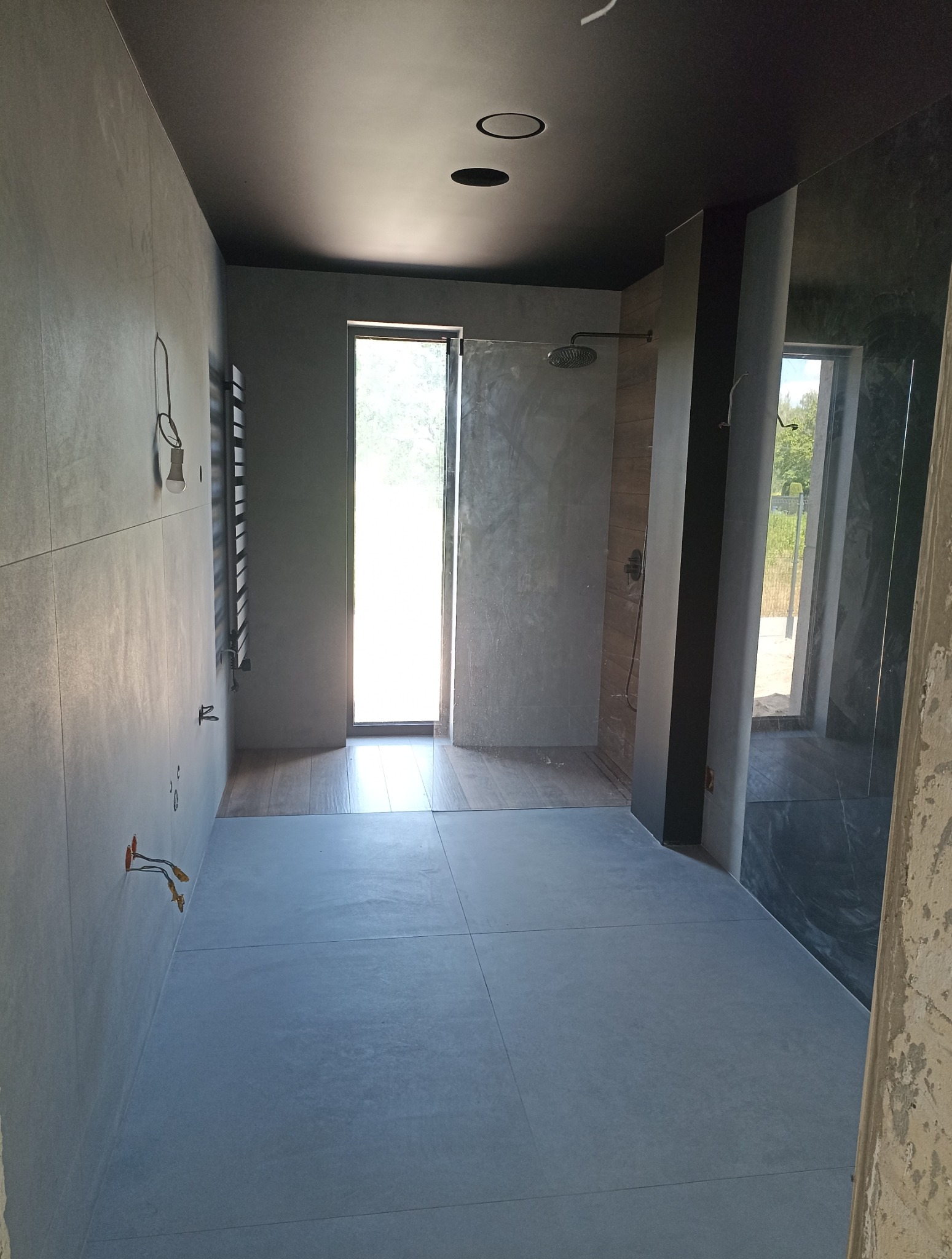 Nowoczesna łazienka w szarościach z czarnym sufitem i punktowym oświetleniem. Widoczne płytki na ścianach i podłodze oraz elementy instalacji.