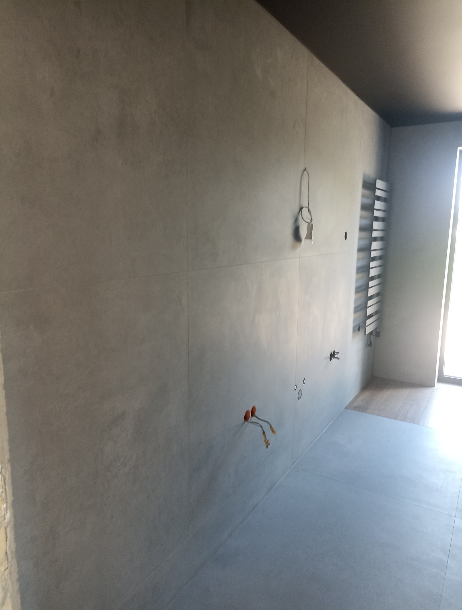 Wykończona łazienka z szarymi płytkami wielkoformatowymi na ścianach i podłodze. Widoczne wystające kable elektryczne i grzejnik drabinkowy. Minimalistyczny design.