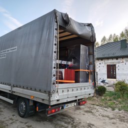 Kuba Iwanowski KK Trans - Ciężarówka z otwartą plandeką, załadowana meblami, stoi na podwórku przed domem w trakcie przeprowadzki. Widoczny numer telefonu na plandece.