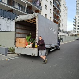 Kuba Iwanowski KK Trans - Otwarta paka busa z kartonami i roślinami, mężczyzna oparty o bok pojazdu na tle nowoczesnego bloku mieszkalnego. Widoczna rampa załadunkowa.