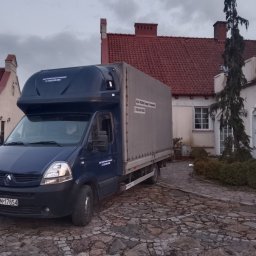 Kuba Iwanowski KK Trans - Niebieski bus transportowy K&K Trans zaparkowany na brukowanym podjeździe przed budynkiem z czerwoną dachówką. Na busie widoczne dane kontaktowe firmy.